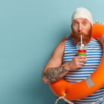 Embarrassed holiday maker drinks cold summer cocktail, spends free time at beach, wears swimcap sailor t shirt, swims with lifebuoy, has surprised expression, models over blue wall with free space